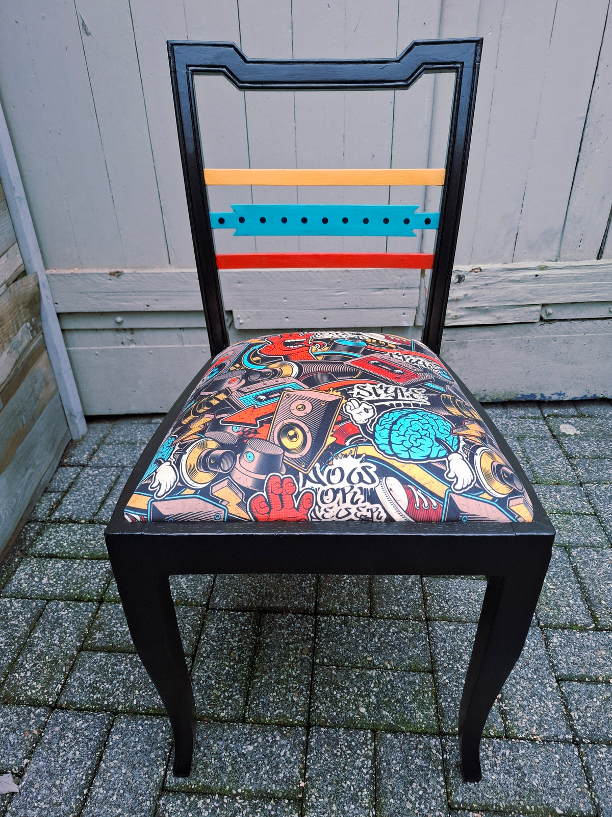 Alter Stuhl komplett aufgearbeitet, schwarzes Holzgestell mit gelben, roten und türkisen Akzenten auf der Rückenlehne, Sitz mit Veloursstoff bezogen mit einem Graffitimuster, obere Frontalansicht Sitz und Rückenlehne