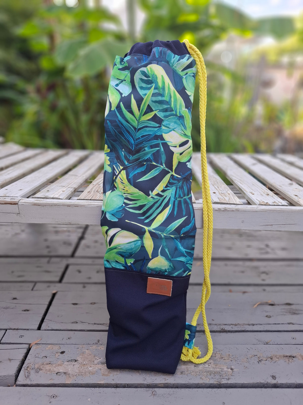 Kellentasche für Saunakellen, oben blau-grünes Muster mit Monstera und anderen Dschungelblättern, unten marineblauer Stoff mit einem Things Outside The Box Lederetikett. Gelbe Kordel. Vorderansicht