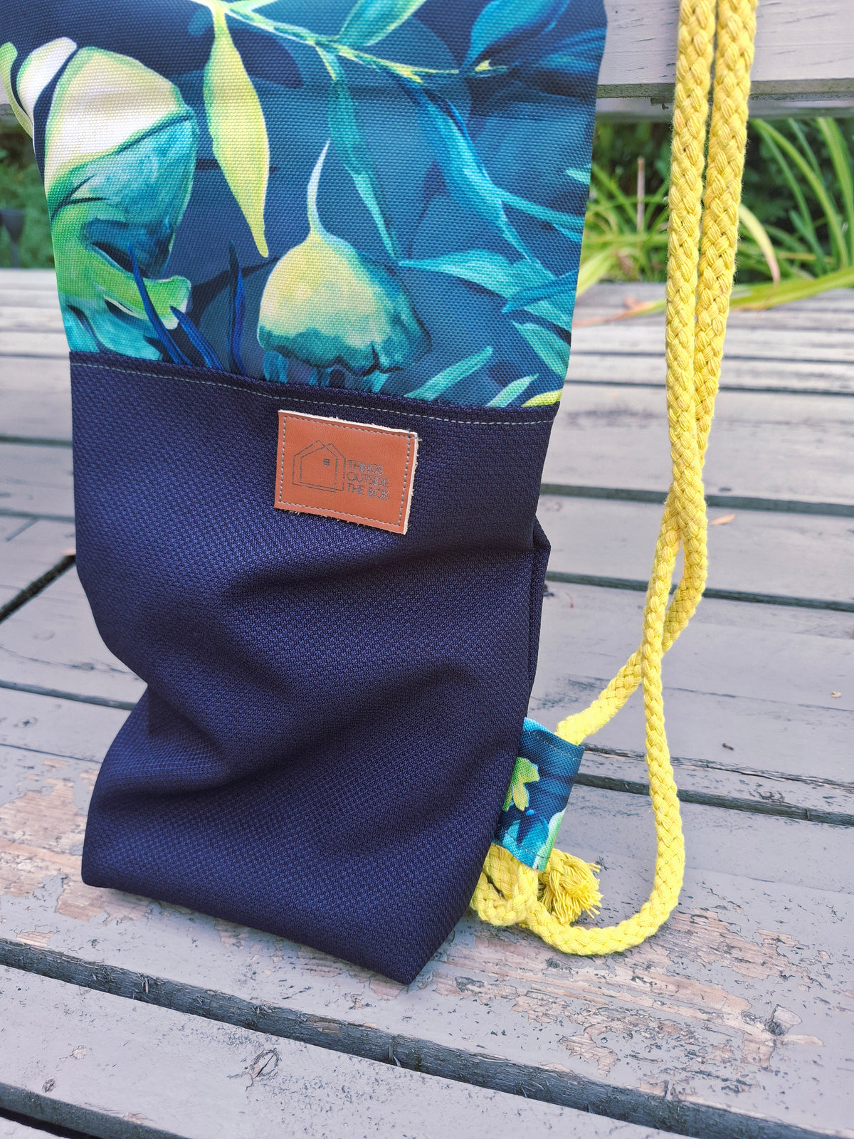 Kellentasche für Saunakellen, oben blau-grünes Muster mit Monstera und anderen Dschungelblättern, unten marineblauer Stoff mit einem Things Outside The Box Lederetikett. Gelbe Kordel. Detailansicht Logoetikett und Kordel