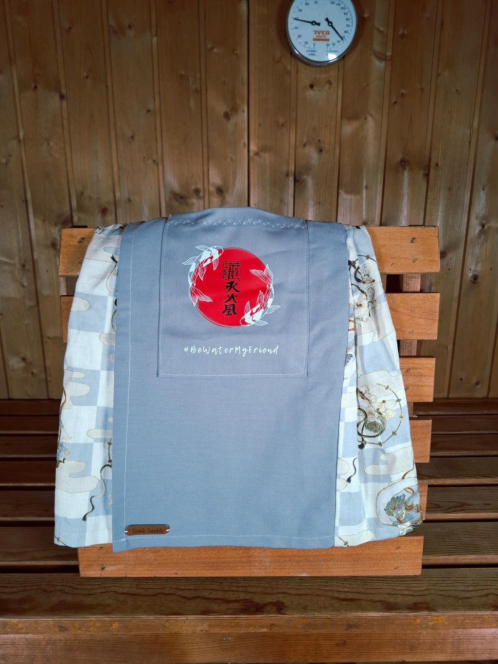 Hanzo Aufguss Art Kilt für Saunameister, japanisches Raijin&Fujin Muster beige-grau, vorne ein hellgrauer Streifen mit einer kleinen Tasche drauf, auf der Tasche das Hanzo Logo, unten ein kleines Logoetikett von Tiröwiki, Vorderansicht Tasche mit Logo