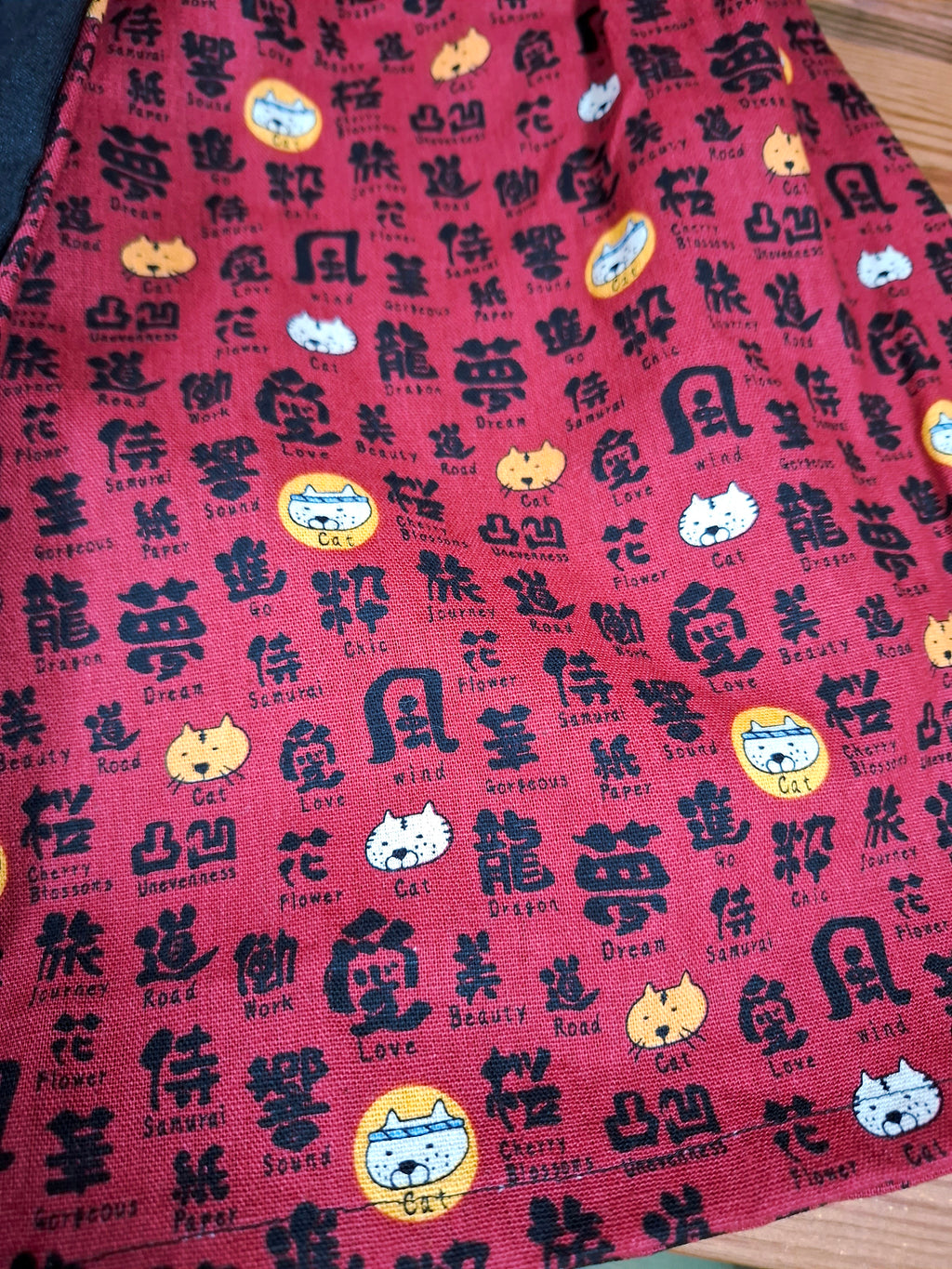 Hanzo Aufguss Art Kilt für Saunameister, japanisches Muster mit Katzen und Kanji auf rotem Hintergrund, vorne ein schwarzer Streifen mit einer kleinen Tasche drauf, auf der Tasche das Hanzo Logo, unten ein kleines Logoetikett von Tiröwiki, Detailansicht Stoff