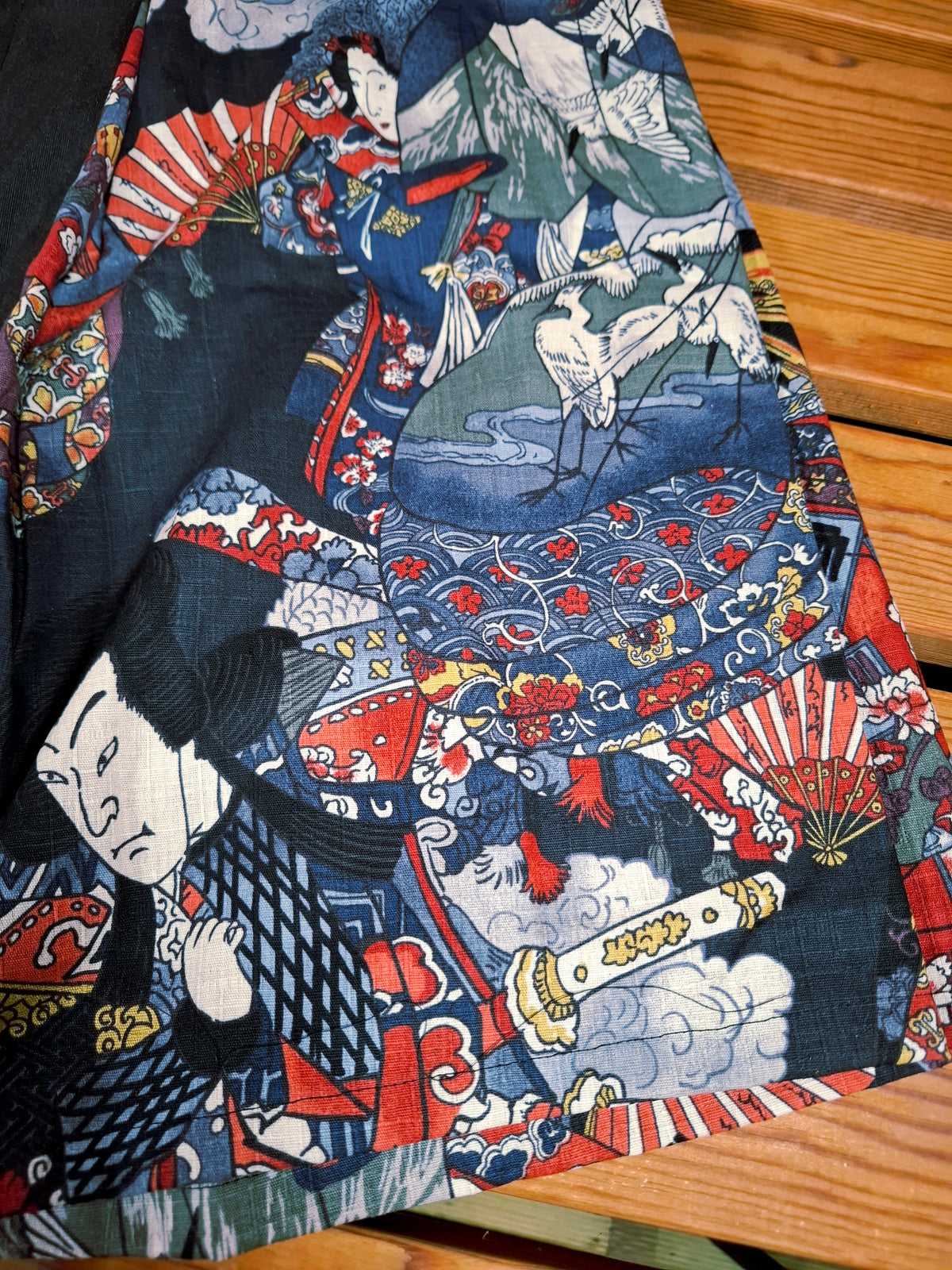Hanzo Aufguss Art Kilt für Saunameister, japanisches Muster mit Samuraj, Kranichen und Fächern, vorne ein schwarzer Streifen mit einer kleinen Tasche drauf, auf der Tasche das Hanzo Logo, unten ein kleines Logoetikett von Tiröwiki, Detailansicht Stoff