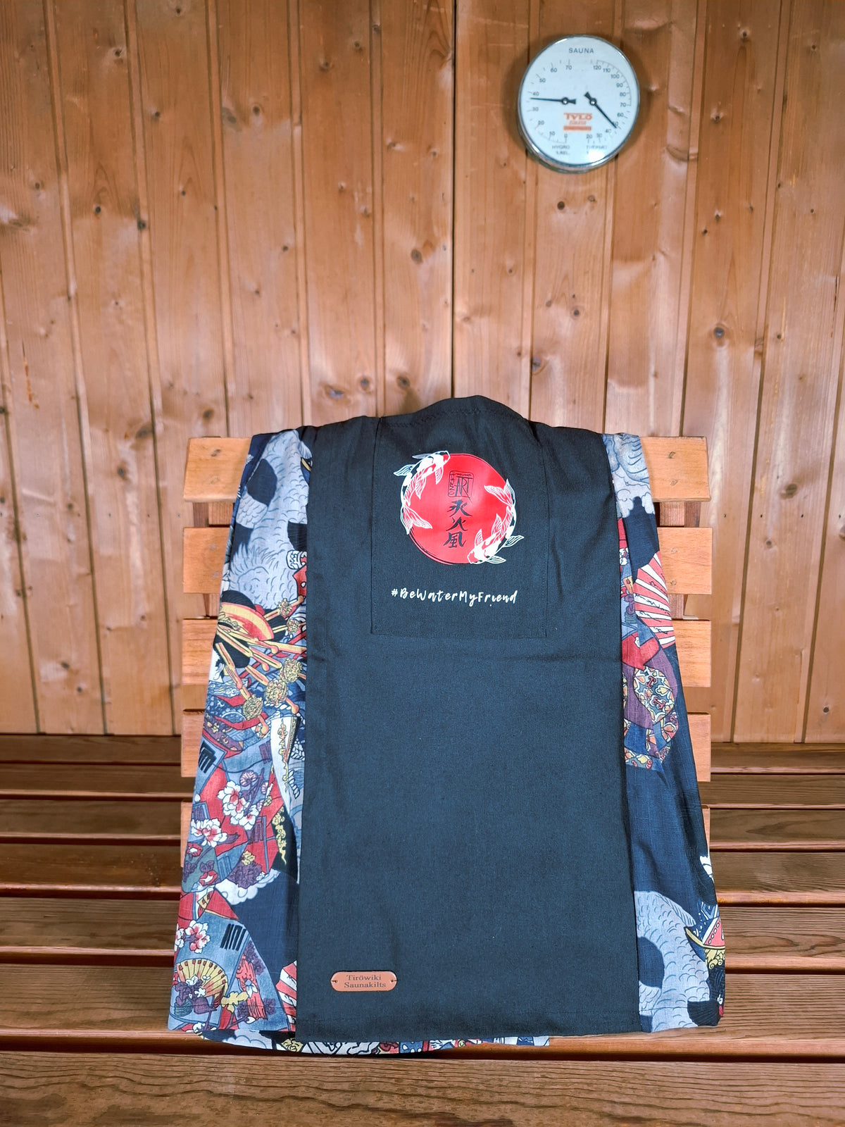 Hanzo Aufguss Art Kilt für Saunameister, japanisches Muster mit Samuraj, Kranichen und Fächern, vorne ein schwarzer Streifen mit einer kleinen Tasche drauf, auf der Tasche das Hanzo Logo, unten ein kleines Logoetikett von Tiröwiki, Vorderansicht Tasche mit Logo