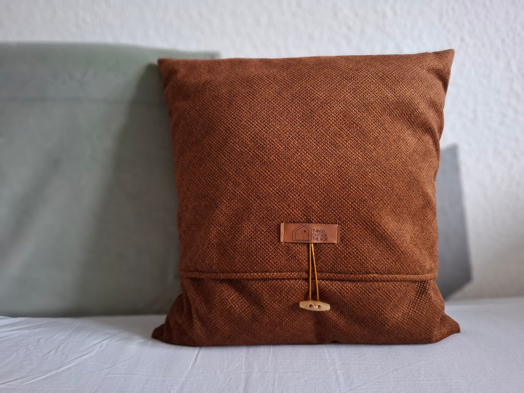 Dekokissen aus braunem Möbelstoff, vorne braunes Logoetikett aus Kunstleder und ein beiger Knopf mit einer goldenen Schleife