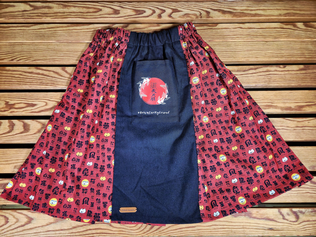 Hanzo Aufguss Art Kilt für Saunameister, japanisches Muster mit Katzen und Kanji auf rotem Hintergrund, vorne ein schwarzer Streifen mit einer kleinen Tasche drauf, auf der Tasche das Hanzo Logo, unten ein kleines Logoetikett von Tiröwiki, Vorderansicht Gesamtansicht